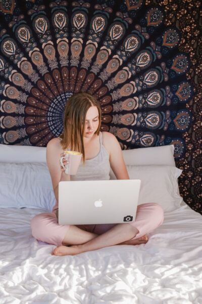 woman sitting on bed using laptop while holding mug